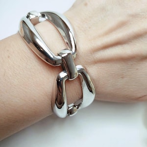 Könnte beinhalten: Ein silbernes Gliederarmband mit großen, ovalen Gliedern. Das Armband befindet sich an einem Handgelenk.