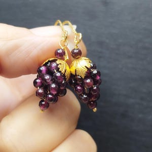 May include: A pair of gold-colored earrings shaped like grape clusters. Each earring features a cluster of small, dark purple beads and gold-colored leaf accents. The earrings are suspended from delicate gold-colored hooks.
