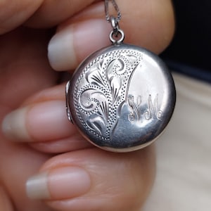 Puede incluir: Un colgante de medallón plateado con un intrincado diseño floral y las iniciales "N M". El medallón redondo cuelga de una delicada cadena de plata. El medallón está sostenido en una mano.