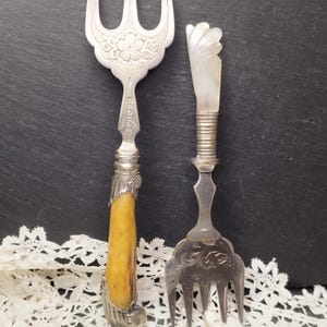 May include: Two antique serving forks. One fork has a silver head with floral detailing and a yellow handle. The other fork has a silver head and a white handle. Both forks are on a black surface with white lace.