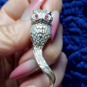 May include: A silver owl pendant with red gemstone eyes. The owl features detailed feathering and a curved, stylized tail. The pendant is likely made of metal and is designed to be worn as jewellery.