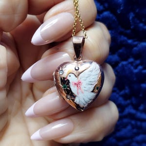 May include: A heart-shaped pendant necklace with a gold chain. The pendant features a painted white dove with a pink bow. The pendant is accented with small gemstones. The necklace is held by a hand with long, manicured nails.