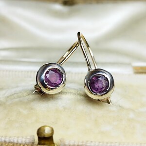 May include: A pair of vintage earrings with a silver setting and gold hooks. Each earring features a round, vivid purple gemstone. The earrings are presented in a cream-coloured jewellery box.