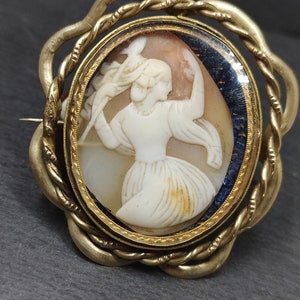 Puede incluir: Un broche de camafeo dorado con un camafeo blanco de una mujer con un vestido fluido. El camafeo está engastado en un marco ovalado dorado con un fondo de esmalte azul. El marco está rodeado por un diseño de cuerda trenzada dorada.