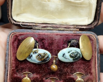 May include: A set of cufflinks in a velvet-lined box. The cufflinks feature a pair of oval-shaped, gold-toned cufflinks with painted images of a dog's head. The box is labeled "Carley, 46 North Michigan Ave., New York, N.Y."
