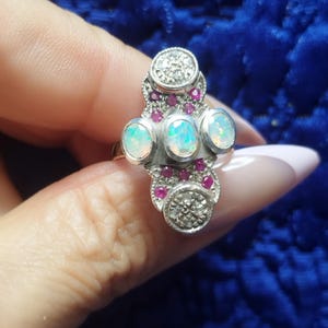 May include: A silver ring with three oval opals and small red gemstones. The ring has a decorative design with two circular sections adorned with small clear stones. The ring is held up by a person's hand.