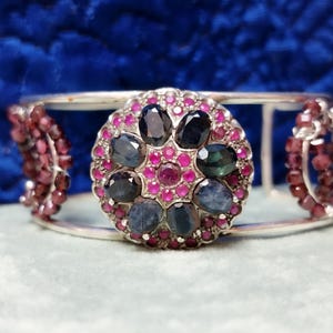 Könnte beinhalten: Ein silberner Armreif mit einem zentralen kreisförmigen Medaillon, das mit ovalen dunkelblauen Saphiren und kleinen runden rosa Rubinen besetzt ist. Die Seiten des Armreifs sind mit kreisförmigen Clustern aus kleinen, dunkelroten Granatperlen verziert.