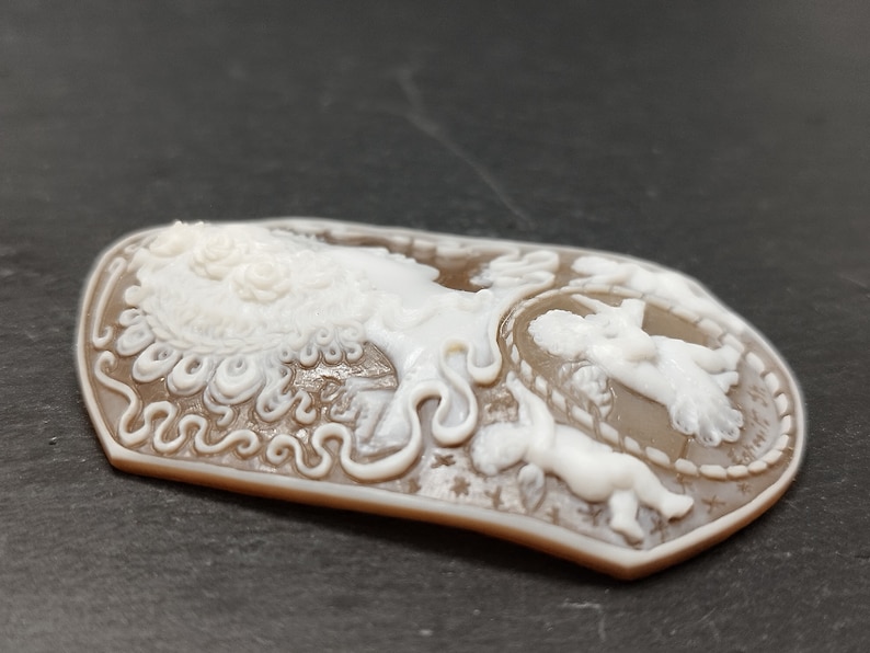 May include: A brown cameo pendant featuring a white carved design of a woman with a crown of flowers and two cherubs. The cameo is set in a silver tone frame.