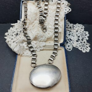 Puede incluir: Un collar plateado con un gran colgante ovalado en una caja azul. La cadena tiene un diseño detallado y enlazado. El colgante es liso con un borde decorativo. Se ve encaje blanco.