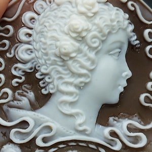 May include: A cameo featuring a woman's profile with curly hair and a floral crown. The cameo is carved from white shell and set in a brown shell background.