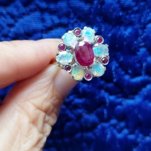 May include: A silver-toned ring featuring a central oval-cut deep red ruby surrounded by a cluster of six round iridescent white opals and six small round red rubies set in a floral pattern, creating a multi-stone halo design on a metal band.
