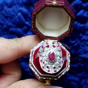 Puede incluir: Un anillo con un rubí central ovalado y diamantes y rubíes circundantes, presentado en un joyero burdeos abierto. El joyero tiene un interior de terciopelo rojo y un cierre dorado. Joyería fina.
