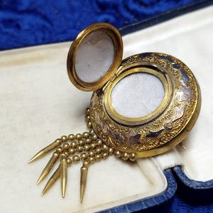 May include: A gold-toned, round locket with an open lid, revealing a white interior. The locket features intricate floral and star designs, with a beaded chain and pointed dangles. It rests in a blue and white box.