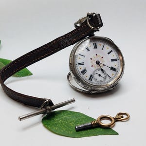 Puede incluir: Un reloj de bolsillo de plata con una esfera blanca y marcadores de hora romanos negros. El reloj tiene una correa de cuero marrón y una llave dorada.