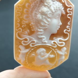 May include: A cameo pendant carved from shell with a detailed design of a woman with flowers in her hair and two cherubs. The cameo is set in a gold-toned metal frame.