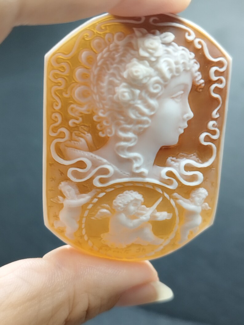 May include: A cameo pendant carved from shell with a detailed design of a woman with flowers in her hair and two cherubs. The cameo is set in a gold-toned metal frame.