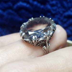 May include: A large, oval-shaped ring with a clear, faceted gemstone set in an ornate silver-toned metal band. The ring's design features intricate filigree details around the stone and on the band. The background is a dark blue.