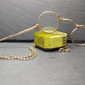 Op de afbeelding: Een antieke monocle in goudkleur met een ketting, rustend op een kleine, vierkante, limoengroene fluwelen doos. De monocle heeft een ronde lens en een handvat met een ring. De ketting is over de doos gedrapeerd.