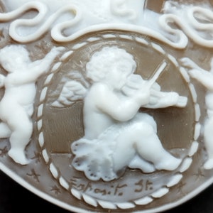 May include: A cameo featuring a cherub playing a violin, surrounded by a rope-like border. The cameo is made of white and brown shell and is signed "Egnosit. St."