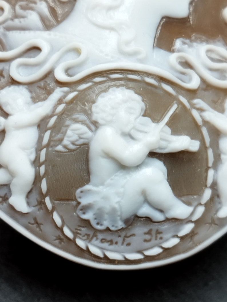 May include: A cameo featuring a cherub playing a violin, surrounded by a rope-like border. The cameo is made of white and brown shell and is signed "Egnosit. St."