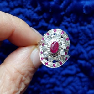 Peut inclure: Une bague en argent ornée d'un rubis ovale central entouré de petites pierres blanches et rouges. La bague présente un motif décoratif en spirale avec un groupe de pierres autour du rubis. La bague est tenue sur un fond bleu.