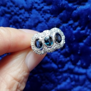 Puede incluir: Un anillo de plata con tres piedras preciosas ovales de color azul oscuro, rodeadas de piedras más pequeñas y transparentes. El anillo se muestra sobre un fondo azul real texturizado, resaltando su diseño detallado y brillo.