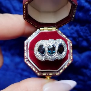 Può includere: Un anello ovale con tre pietre preziose blu scuro, circondate da pietre più piccole e chiare. L'anello è presentato in un portagioie bordeaux con interno in velluto rosso. La scatola ha un bordo dorato.