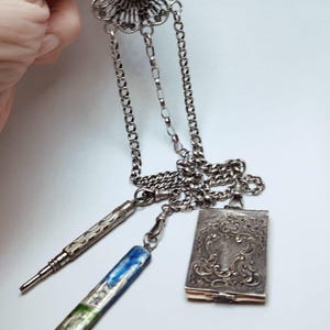 Puede incluir: Un collar colgante antiguo plateado con un diseño floral de filigrana y cadenas colgantes. El collar incluye un lápiz, un bolígrafo con detalles azules y verdes y un pequeño colgante ornamental en forma de libro.