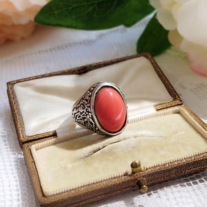 Op de afbeelding: Een zilveren ring met een ovale koraalsteen gezet in een gedetailleerd bloemmotief. De ring wordt gepresenteerd in een bruine fluwelen ringdoos.