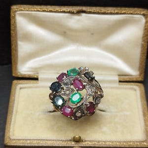 May include: A vintage ring featuring a cluster of gemstones in shades of green, red, and blue, set in a silver filigree design. The ring is presented in a vintage jewellery box with a cream satin lining.