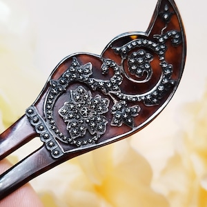 Puede incluir: Un peine para el cabello de carey marrón con detalles de filigrana plateada. El peine tiene dos púas y un diseño decorativo de una flor y hojas.