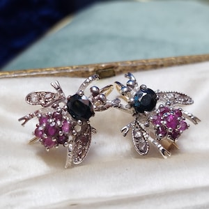 May include: A pair of silver bee earrings. Each earring has a dark blue gemstone head, a cluster of small, round, magenta gemstones, and detailed wings. The earrings are presented on a white surface within a gold-edged case.
