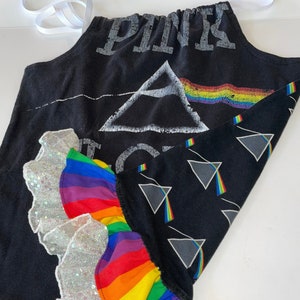 May include: Black apron with a rainbow-colored ruffle design and a silver sequined trim. The apron features the text "PINK" and a graphic of a prism with a rainbow. White ribbon ties are attached.