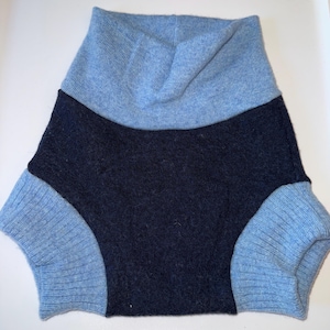 May include: A pair of knit shorts featuring a dark navy blue centre panel and a light blue waistband and leg cuffs. The garment is constructed from a soft, textured knit fabric with a ribbed pattern on the light blue sections and a solid knit.