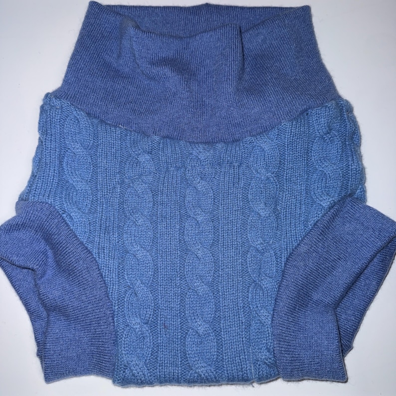 May include: A light blue knit garment featuring a high turtleneck collar and a central cable knit pattern. The piece has ribbed cuffs at the leg openings and a smooth, solid-coloured knit waistband, constructed from a soft, textured blue yarn.