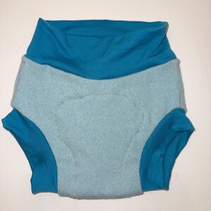 May include: A light blue and teal cloth diaper with a high waist and leg openings. The diaper is made of a soft, textured material. The waist and leg openings are teal, while the main body is light blue.