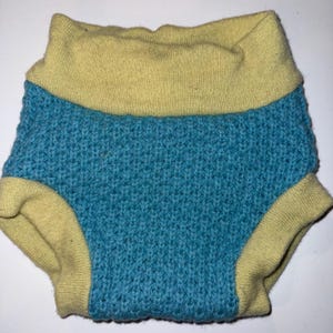 May include: A knitted baby diaper cover in a teal blue color with a yellow waistband and leg openings. The diaper cover is made of a textured knit fabric.