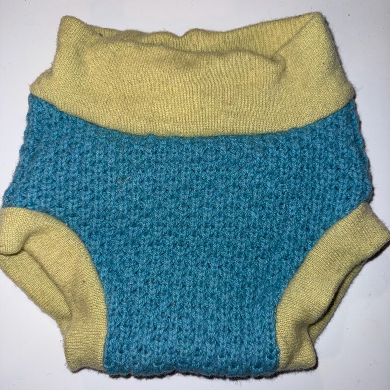 May include: A knitted baby diaper cover in a teal blue color with a yellow waistband and leg openings. The diaper cover is made of a textured knit fabric.