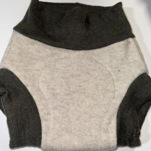 May include: A two-tone knitted garment in light beige and dark grey. The garment appears to be a diaper cover or similar item, with a high waistband and leg openings. The fabric has a soft, textured appearance.