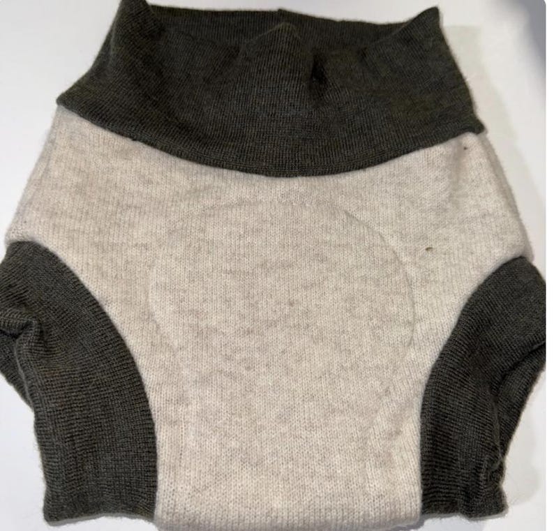 May include: A two-tone knitted garment in light beige and dark grey. The garment appears to be a diaper cover or similar item, with a high waistband and leg openings. The fabric has a soft, textured appearance.