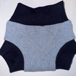 May include: A light blue sweater with a V-neck design and navy blue accents. The sweater has navy blue sleeves and a navy blue collar. The sweater is folded, showing the front and part of the sleeves.