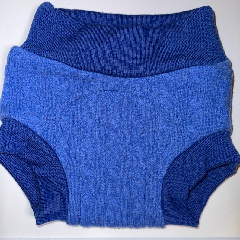 May include: A pair of pull-on nappy covers in a vibrant blue shade. The garment features a wide, ribbed waistband and leg cuffs in a darker blue tone, while the main body displays a textured cable knit pattern with a reinforced central panel.