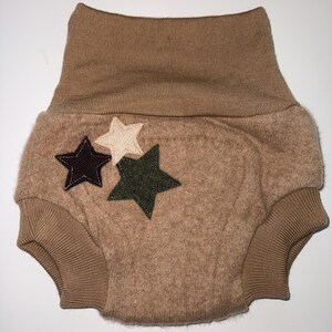 May include: A tan baby nappy cover with a high waistband and ribbed leg openings. Three star appliqu&eacute;s in dark purple, cream, and olive green are sewn onto the front. The nappy cover is made of a soft, textured fabric.