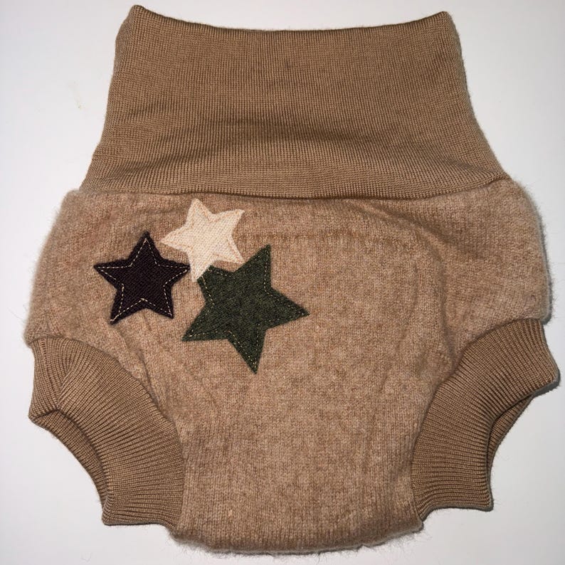 May include: A tan baby nappy cover with a high waistband and ribbed leg openings. Three star appliqu&eacute;s in dark purple, cream, and olive green are sewn onto the front. The nappy cover is made of a soft, textured fabric.