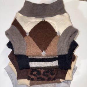 Puede incluir: Una pila de suéteres para perros en varios colores y patrones. El suéter superior presenta un diseño argyle marrón y crema. Otros suéteres incluyen colores lisos, rayas y un estampado de leopardo. Cada suéter tiene un cuello alto.