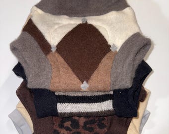 May include: A stack of dog sweaters in various colors and patterns. The top sweater features a brown and cream argyle design. Other sweaters include solid colors, stripes, and a leopard print. Each sweater has a turtleneck collar.