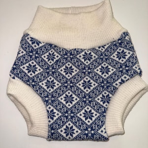 May include: A pair of knitted nappy covers featuring a blue and white diamond pattern with snowflake motifs. The garment has a wide, ribbed cream-coloured waistband and matching ribbed leg openings, constructed from a soft, thick knit fabric.