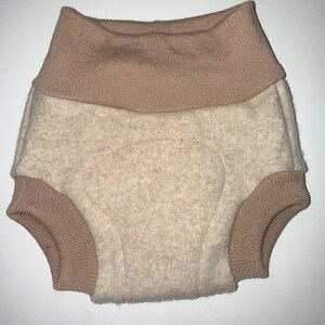 May include: A pair of beige baby shorts with a wide, ribbed, light brown waistband and leg cuffs. The shorts are made of a soft, textured fabric, likely wool or a wool blend. The design is simple and functional.