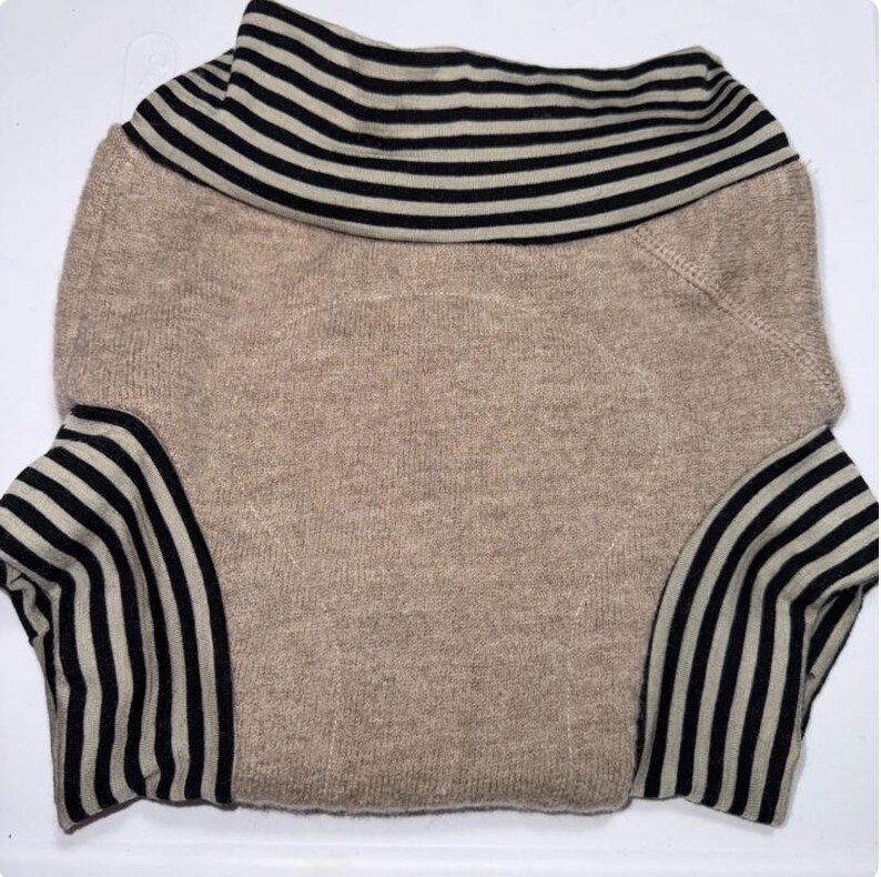 May include: A beige sweater featuring black and white striped detailing on the collar and sleeves. The sweater has a wide, folded collar and long sleeves. The design is simple and classic, suitable for casual wear.