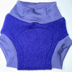May include: A two-tone garment in shades of blue and purple. The main body is a textured, deep blue, while the collar and leg openings are a lighter purple. The garment appears to be made of a soft, knitted material.